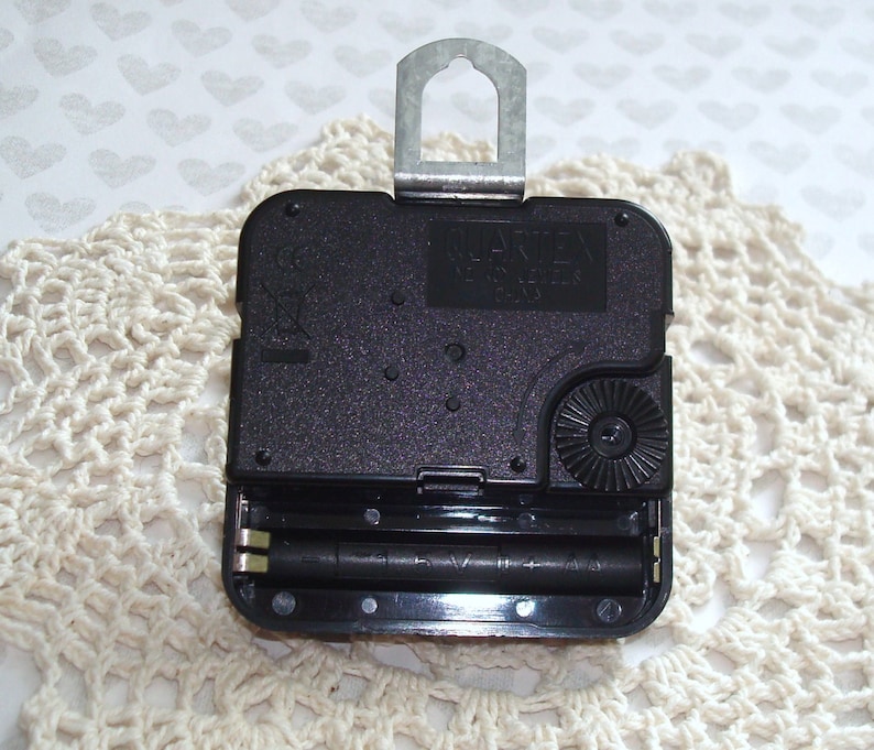 May include: A black quartz clock movement with a metal hanger. The clock mechanism has a battery compartment holding two AA batteries. The clock movement is on a cream-colored, crocheted doily.