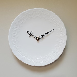 May include: White decorative plate repurposed as a wall clock. The plate has an embossed floral design around the edge. Black clock hands and a brass-colored center complete the design. The clock is mounted on a neutral-colored wall.