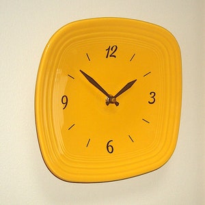 May include: A square, yellow wall clock with black numerals and hands. The clock has a rounded square shape and is mounted on a white wall. The clock face is a solid yellow color.