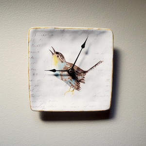 May include: Square wall clock featuring a watercolor illustration of a singing bird. The clock face has a vintage aesthetic with handwritten script and a gold-colored trim. The bird is brown, blue, and yellow.