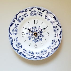 May include: A round wall clock made from a white plate with a blue floral design. The clock face features black numerals and hands, with a gold-colored center. The plate has a decorative blue border.
