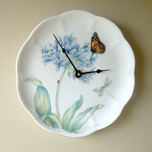May include: A white porcelain plate with a blue flower and green leaves design. A butterfly and dragonfly are also depicted on the plate. The plate has a clock face with black hands.