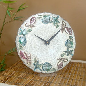 May include: A round, off-white clock with a textured surface, adorned with seashells and starfish in shades of blue, green, and pink. The clock hands are black. The clock is a decorative item with a coastal theme.