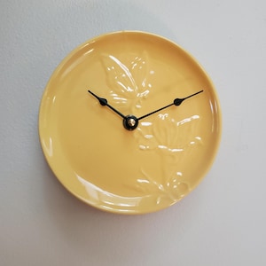 May include: A yellow ceramic plate repurposed as a wall clock. The clock features black hands and a black center, with a raised floral design on the plate's surface. The clock is mounted on a light gray wall.