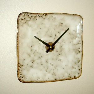 May include: A white ceramic square clock with black hands and a brown center. The clock has a speckled, textured surface.