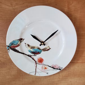 May include: White ceramic plate clock featuring two birds perched on a branch with red and pink flowers. The birds have blue, brown, and red accents. Black clock hands and a gold center complete the design. The clock is on a wooden surface.