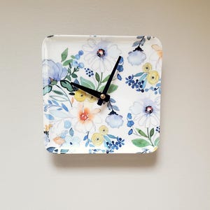 May include: A white square clock with black hands and a floral design featuring blue, yellow, and green watercolor flowers.
