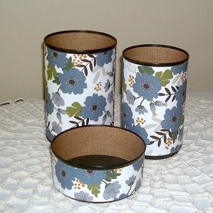 May include: Three cylindrical containers with a brown top and a white background with a blue floral pattern. The containers are on a white crocheted surface.