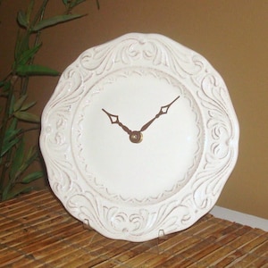 May include: White ceramic clock with ornate detailing on the face. The clock has gold-colored hands.