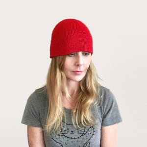 May include: A bright red knit beanie hat. The hat is a solid color and appears to be made of a soft, ribbed knit material. The beanie is worn on the head. The person in the image is wearing a gray t-shirt.