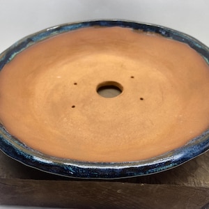May include: A terracotta colored ceramic pot with a blue and green glaze around the rim. The pot has a drainage hole in the center and four smaller drainage holes around the rim.