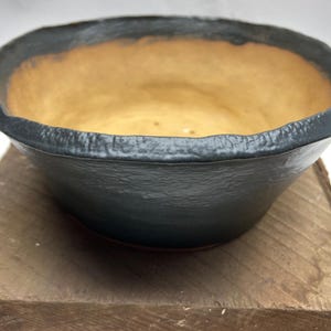 May include: A dark gray ceramic pot with a light brown interior. The pot has a rounded shape and a slightly uneven rim.