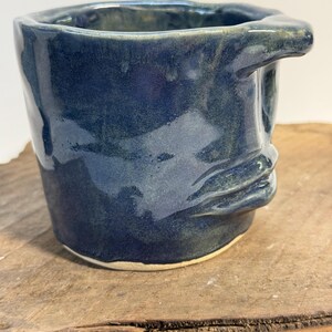 May include: A handmade ceramic mug with a dark blue glaze. The mug has a unique, organic shape with a handle and a textured surface. The mug is sitting on a wooden surface.