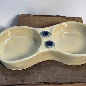 May include: A light yellow ceramic double dish with two blue glazed accents. The dish is slightly curved and has a unique shape.