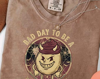 May include: Espresso-colored t-shirt with a graphic of a deviled egg character with horns, a sinister grin, and the text "BAD DAY TO BE A DEVIELED EGG." A silver necklace is draped across the shirt.