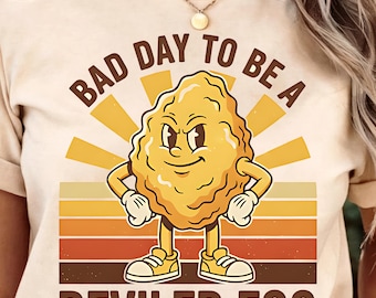 May include: Beige t-shirt with a graphic of a deviled egg character with the text "BAD DAY TO BE A DEVILED EGG". The design features a cartoon deviled egg with arms, legs, and a determined expression, set against a retro sunset.