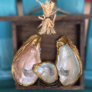 Può includere: Un presepe decorativo realizzato con conchiglie di ostrica. Le conchiglie sono dipinte con bordi dorati e colori tenui, disposte su una base di legno. Un fascio di paglia è appeso sopra la scena, aggiungendo un tocco rustico. Lo sfondo è blu.