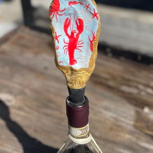 Könnte beinhalten: Ein goldfarbener Flaschenverschluss aus Austernschalen mit einem roten Hummer-Design. Der Verschluss befindet sich auf einer Flasche Rotwein mit der Aufschrift "Villa San Giovanni Chianti".