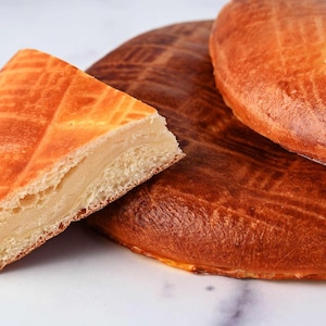 May include: A close-up of a slice of golden-brown pastry with a creamy filling, next to two whole pastries. The pastries have a lattice pattern on top. The baked goods are on a white marble surface.