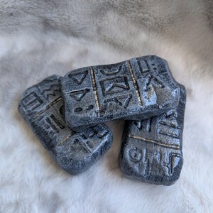 May include: Three rectangular, grey stone tablets with gold-coloured accents and etched symbols. The tablets are arranged on a white, fluffy surface. The stone tablets have a weathered appearance.