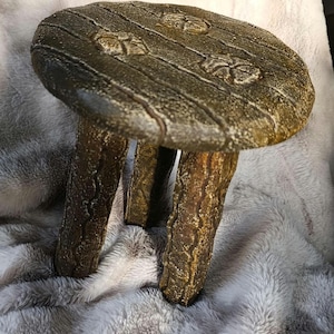 以下が含まれることがあります： 丸い天板と3本の頑丈な脚が付いた、小さくて素朴な木製のスツール。 天板は濃い茶色で、木目調のテクスチャと装飾的な花のアクセントがあります。 脚は木の枝のように見えるようにテクスチャ加工されています。