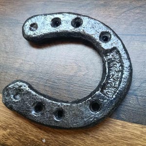 Op de afbeelding: Een zilveren en zwart hoefijzer met zichtbare spijkergaten. Het hoefijzer is gemaakt van een textuurrijk materiaal en ligt op een houten oppervlak. Het hoefijzer is een traditioneel symbool van geluk.