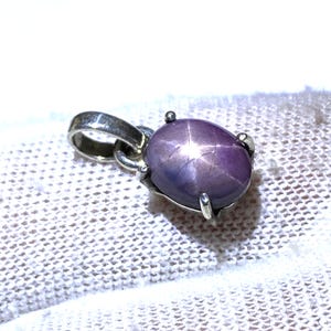 May include: A silver pendant with a large, oval, purplish-pink star sapphire gemstone. The gemstone displays a six-ray star effect. The pendant is set in a silver metal setting with a loop for a chain.