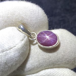 May include: A silver pendant with an oval, purplish-red gemstone. The gemstone displays a star-like pattern. The pendant has a simple, elegant design. The pendant is suitable for everyday wear or special occasions.