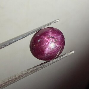 Può includere: Una gemma cabochon di forma ovale color magenta intenso, con una superficie liscia e lucida e inclusioni interne visibili, tenuta saldamente tra le due punte metalliche di una pinzetta color argento su uno sfondo di colore chiaro.