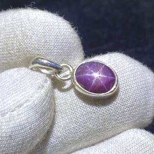 Può includere: Ciondolo ovale con una gemma di rubino stellato. Il rubino è di un colore rosso-violaceo intenso con un motivo a stella a sei raggi. Il ciondolo è incastonato in una montatura in metallo argentato con un anello per una catena.