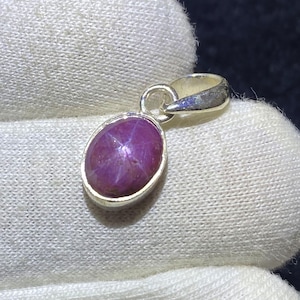 May include: A silver pendant with an oval, deep-red gemstone. The gemstone has a star-like pattern across its surface. The pendant is set in a silver bezel and has a small loop for a chain. The background is a white fabric.