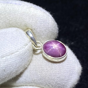 May include: A silver pendant with an oval, purplish-pink star ruby gemstone. The gemstone has a six-rayed star pattern. The pendant is set in a silver bezel and has a silver bail for a chain. The gemstone is approximately 1.5 cm in length.