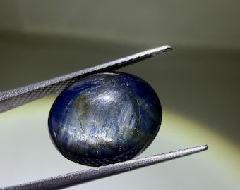 May include: A polished, oval-shaped blue gemstone with visible internal striations and dark inclusions. The stone displays a deep navy color with lighter, reflective streaks across its surface, held securely by a pair of metal tweezers.