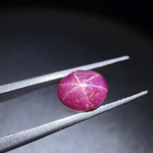 Puede incluir: Una piedra preciosa de rubí estrella de corte cabujón redondo con un tono rojo magenta intenso. La piedra muestra un efecto de estrella de seis rayos, conocido como asterismo, que irradia desde el centro. Está sostenida por unas pinzas de metal plateado.