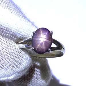 May include: A silver ring featuring a large, oval, star-shaped purple gemstone. The gemstone is set in a four-prong setting, and the ring band has a split design. The ring is shown against a white background.