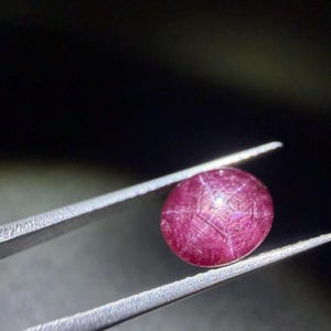 Peut inclure: Une pierre précieuse rubis étoilé de taille cabochon ronde, présentant une teinte rouge magenta profonde. La pierre affiche un effet d'astérisme distinct à six rayons sur sa surface, formé par des lignes lumineuses qui créent une étoile.