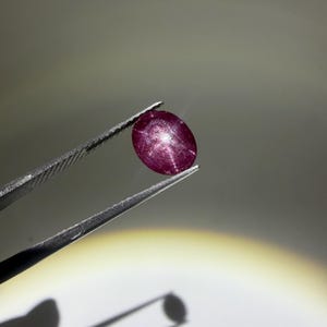 Op de afbeelding: Een ovaalvormige, diep magenta sterrobijn edelsteen met een duidelijk zesstralig stereffect over het oppervlak. De in cabochon geslepen steen vertoont een doorschijnende kwaliteit met zichtbare interne insluitingen en een gladde bovenkant.