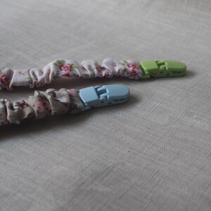 May include: Two fabric baby teethers with plastic clips. One teether is pink with floral print and a green clip. The other teether is brown with a floral print and a blue clip.