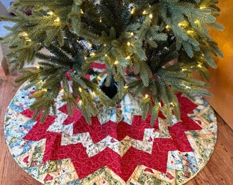 May include: A Christmas tree skirt with a red and white star pattern. The skirt is made of fabric and has a circular shape with a hole in the center for the tree trunk. The skirt is decorated with a patchwork design and is placed on a wooden floor.