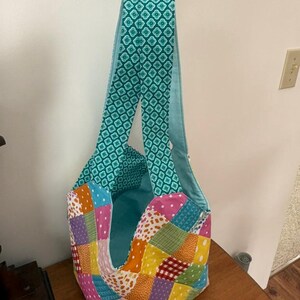 May include: A handmade patchwork tote bag with a teal geometric patterned strap and interior. The bag's exterior features a colorful patchwork design with squares of various colors and patterns, including pink, orange, and green.