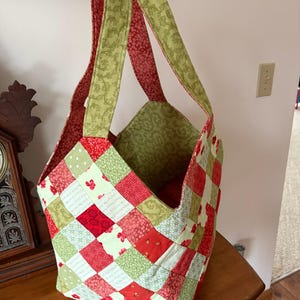 May include: A handmade patchwork tote bag with a quilted design. The bag features a mix of red, green, and white fabric squares. The handles are green with a floral pattern, and the interior is a matching green fabric.