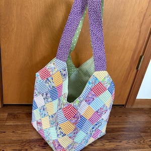 May include: A patchwork tote bag with a variety of colorful squares. The bag has purple straps with a white floral pattern and a green polka dot lining. The bag is open, revealing the interior.