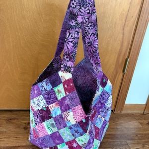 May include: A handmade tote bag with a patchwork design in shades of purple, pink, and blue. The bag features a variety of floral and geometric patterns. The straps are a darker purple with a floral print. The bag is open and appears to be empty.