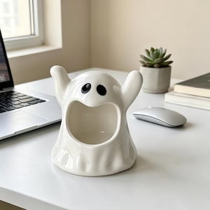 Może przedstawiać: Błyszcząca, biała ceramiczna figurka ducha z dużym, pustym, owalnym otworem pośrodku i dwoma uniesionymi ramionami. Duch ma dwoje prostych, czarnych, owalnych oczu umieszczonych nad szerokimi, otwartymi ustami, tworząc puste w środku naczynie.