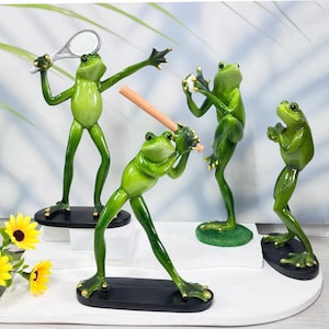 Könnte beinhalten: Vier grüne Froschfiguren, die verschiedene Sportarten spielen. Ein Frosch hält einen Tennisschläger, ein anderer einen Baseballschläger, ein anderer einen Baseball und der letzte Frosch steht mit verschränkten Armen da.