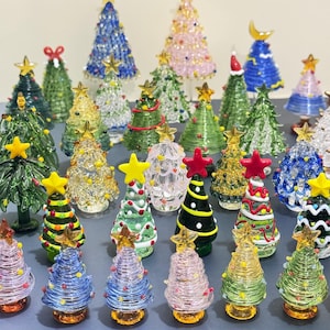 May include: A collection of miniature glass Christmas trees in various colors, including green, blue, pink, and yellow. Each tree is adorned with star toppers and decorative accents. Some trees have a clear, textured appearance, while others are solid.
