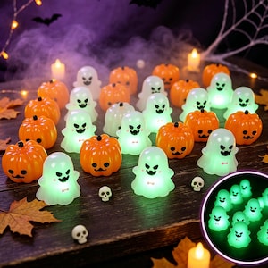 Puede incluir: Una colección de pequeñas figuras LED brillantes con forma de calabazas naranjas y fantasmas blancos con rasgos faciales negros. Las figuras están dispuestas sobre una superficie de madera oscura junto a hojas de otoño y pequeños cráneos.