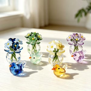 Puede incluir: Cuatro pequeños jarrones de cristal, cada uno con un ramo de flores de cinco pétalos con centros amarillos, junto con cuatro figuras de pájaros de cristal a juego en azul, verde claro, amarillo y morado, dispuestos sobre una superficie plana.
