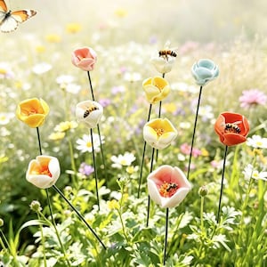 May include: A collection of ceramic flower sculptures on thin metal stems, featuring various pastel and vibrant colours including yellow, pink, white, blue, and orange, with small bee figures attached to several of the petals in a garden setting.