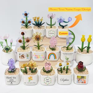 May include: A collection of glass flower sculptures in small white pots. Each pot features a different flower design, including sunflowers, tulips, and daisies. Some pots have custom text or photos, and others have decorative elements like rainbows.